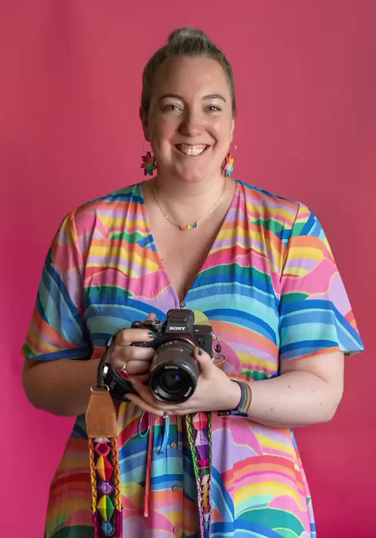 A white female photographer in a rainbow striped dress in front of a pink background, smiling and holding her camera.