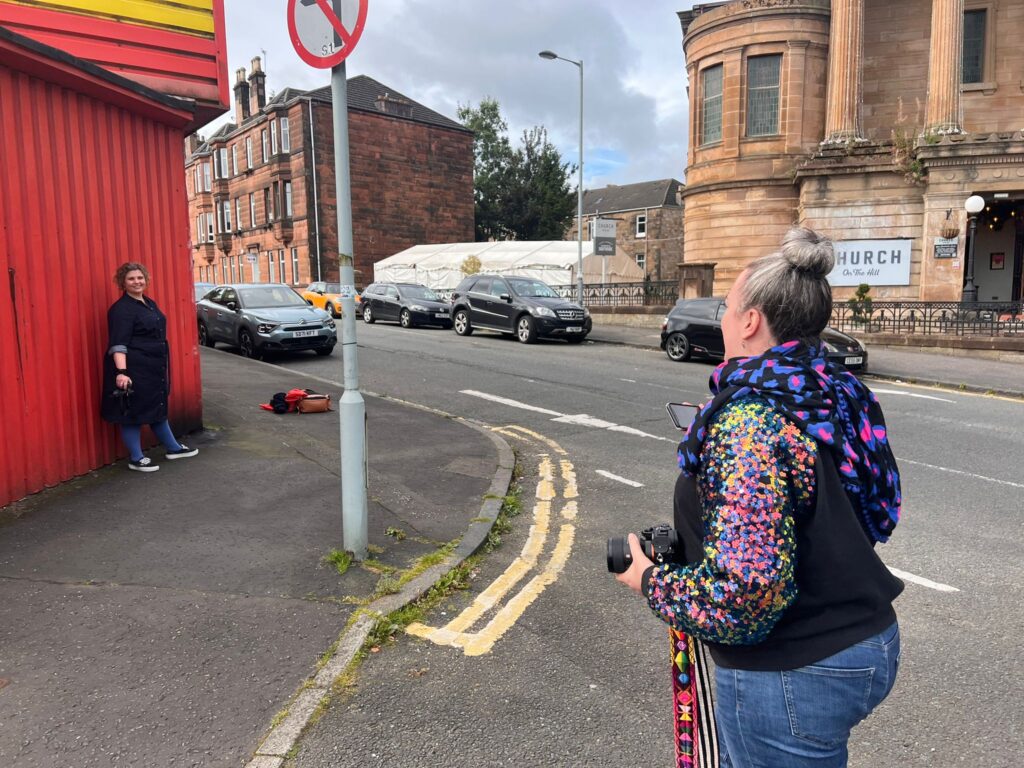 A white female photographer in a sequin sweater directing a  white woman to pose in front of a red wall of an MOT test centre in Glasgow
