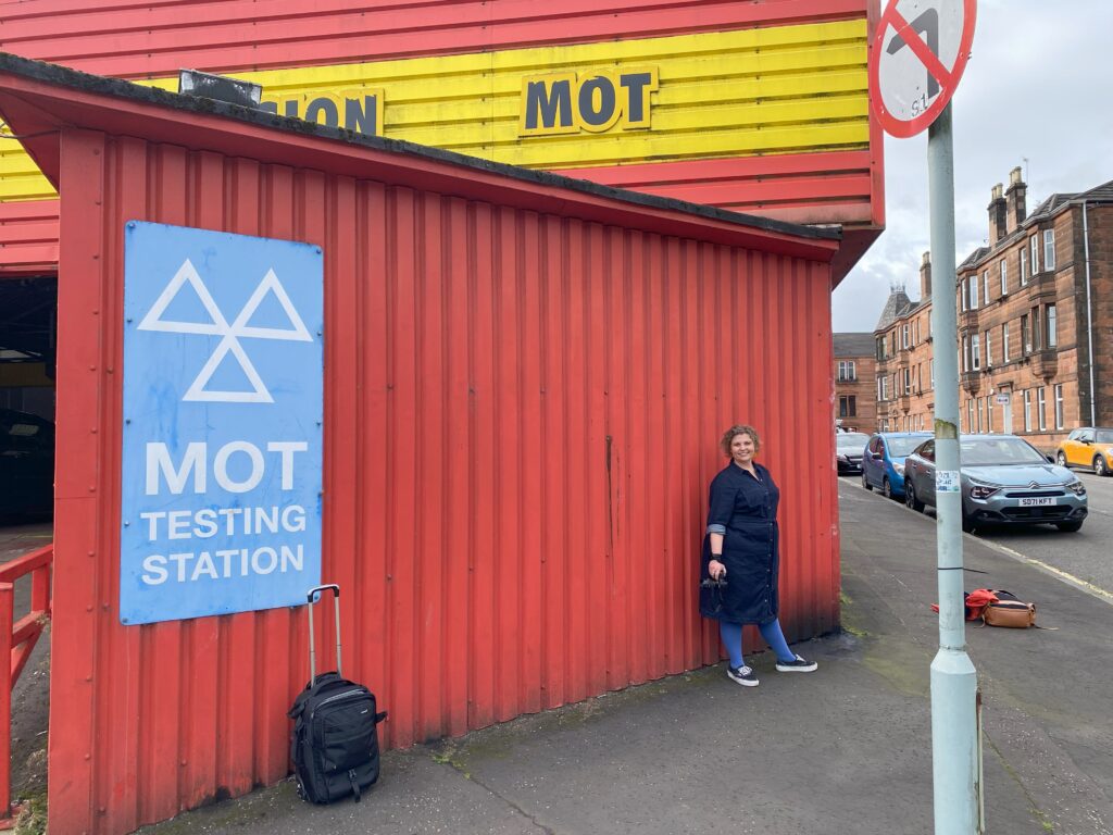 A white woman posing in front of the red wall of an MOT test centre in Glasgow