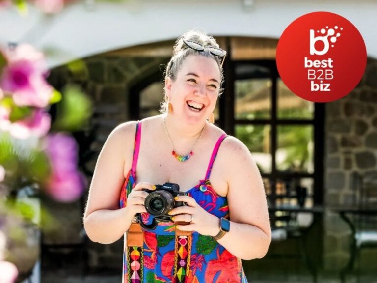 Photo of Bex, a white woman wearing a blue and red jumpsuit in the sunshine. She is holding her camera and smiling. There is a badge in the corner showing that Bex won Best B2B business award in 2024.