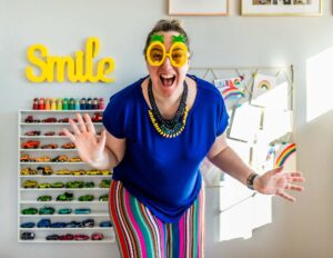 a white woman with brown hair posing in a silly way in front of a yellow sign that says smile, she's wearing pineapple sunglasses and a blue t-shirt with rainbow striped trousers
