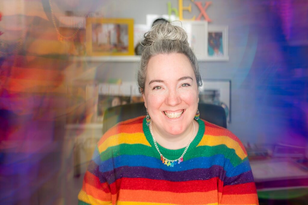 White woman wearing a rainbow striped jumper, looking happy in her office