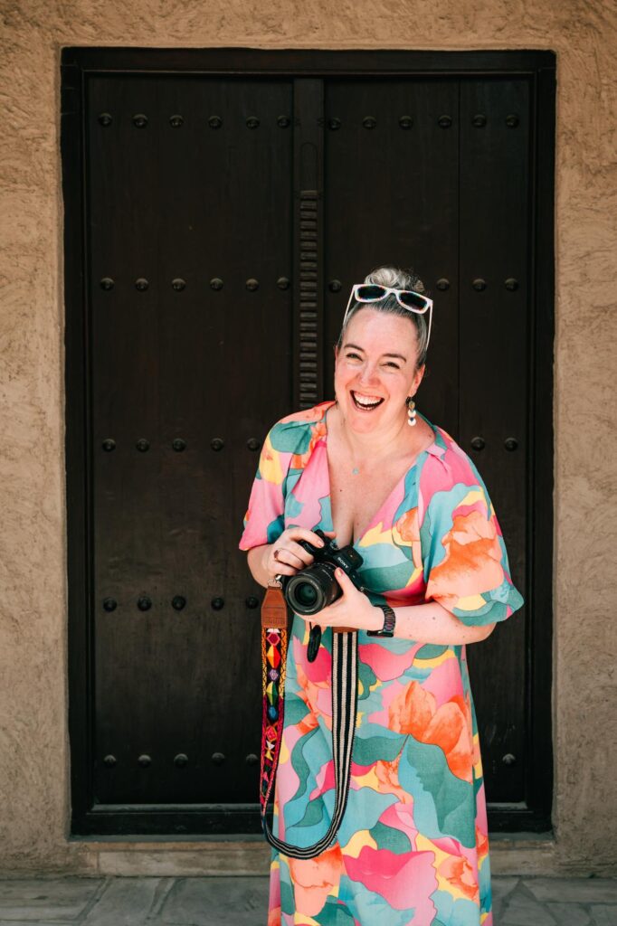 A white woman wearing a multi-coloured dress is holding a camera and laughing, standing in front of a brown door in the sunshine in Dubai