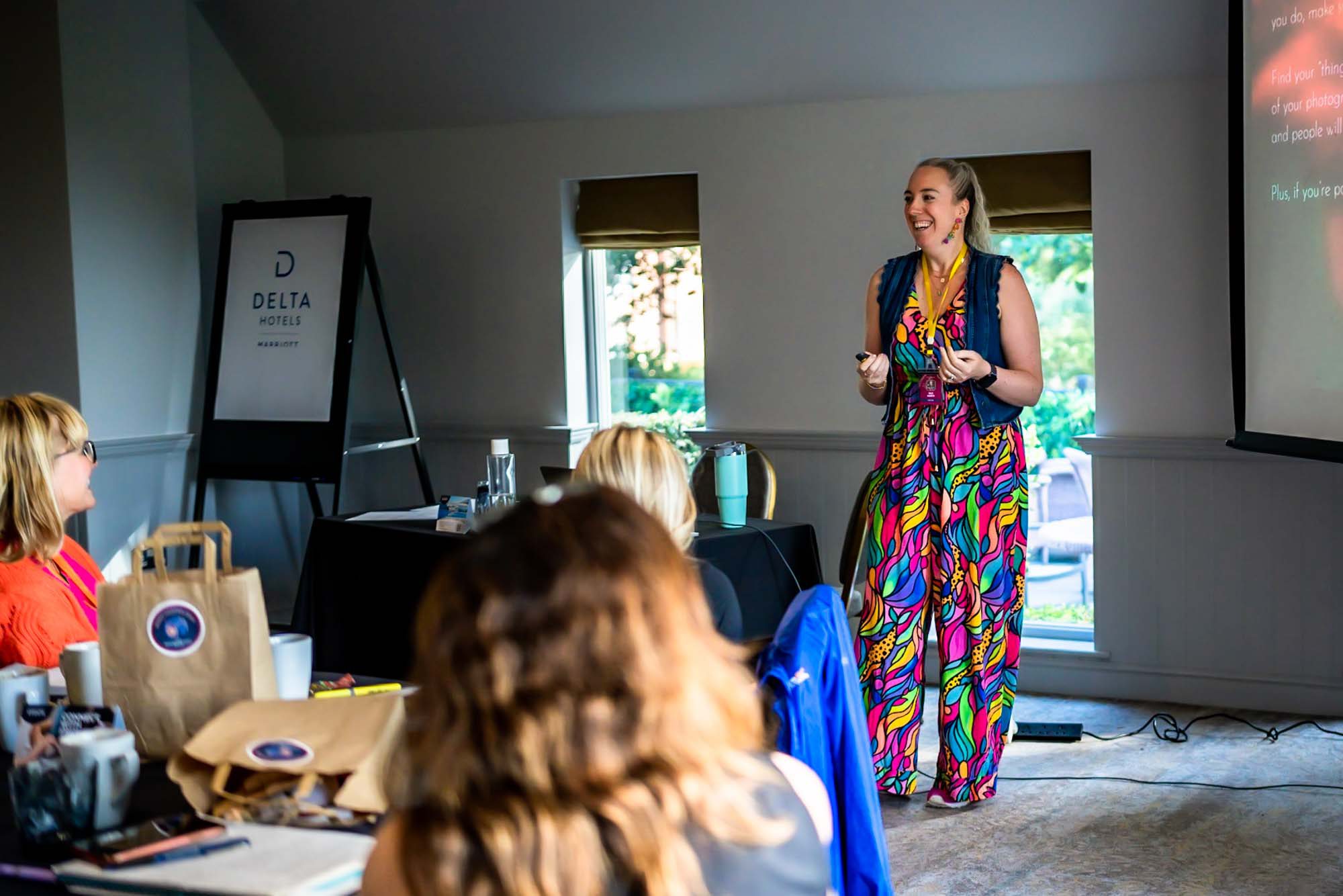 A white woman in a multi-coloured jumpsuit and denim waistcoat is presenting to a room of people at a photography conference.