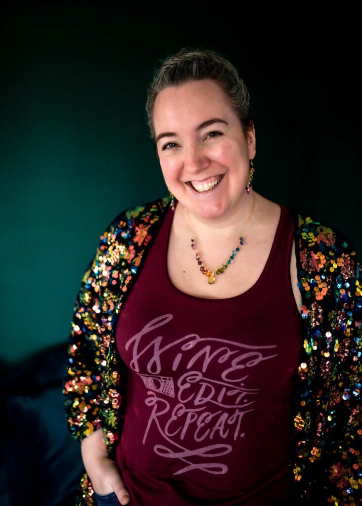 A white woman wearing a vest which says "Wine, Edit, Repeat" and a rainbow necklace, smiling at the camera.
