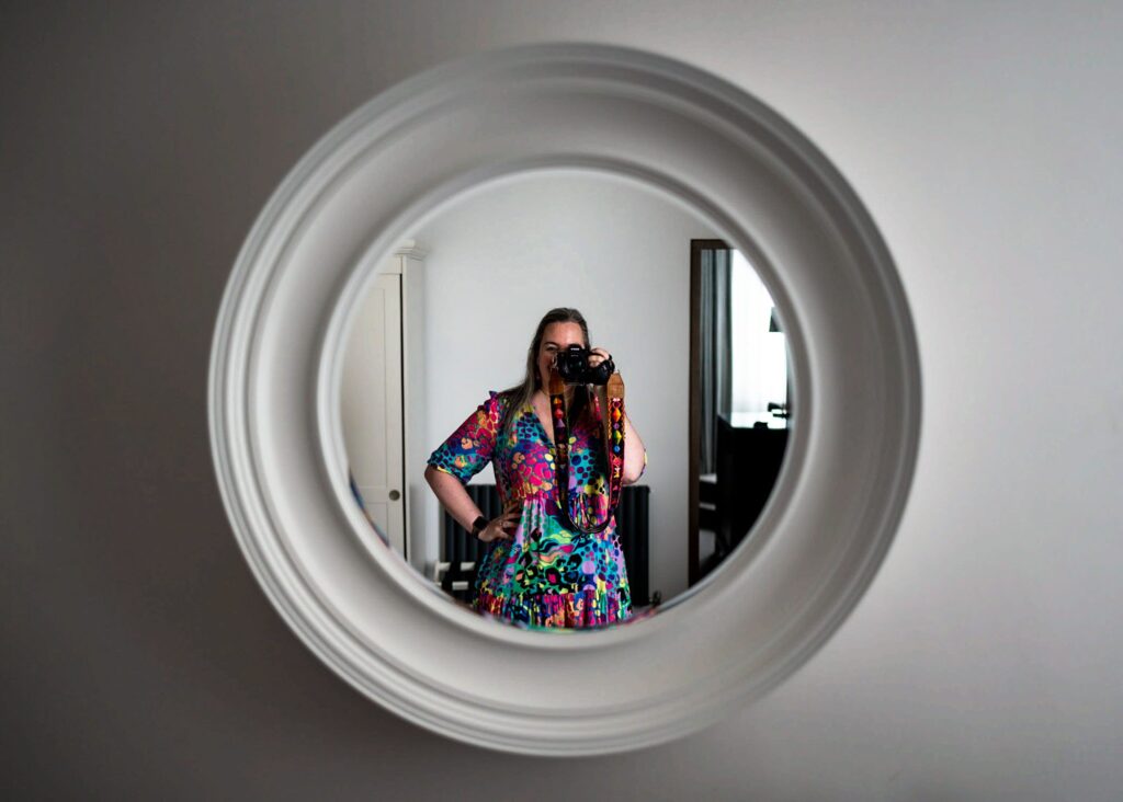 A female photographer wearing a rainbow patterned dress, taking a self-portrait in a round white hotel mirror.