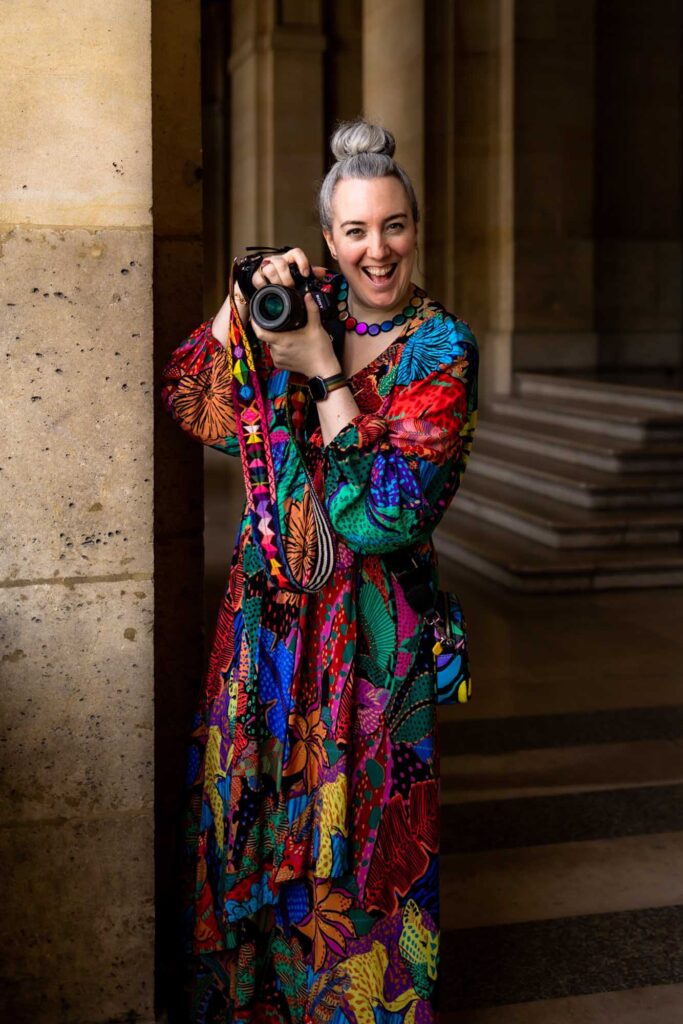 A happy, smiling white woman in a multi-coloured dress is holding her camera up to take photos in Paris