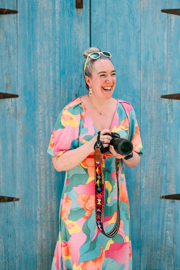 White woman with grey hair in front of a blue door. She is wearing a multi-coloured dress, holding a professional camera and smiling to the side.