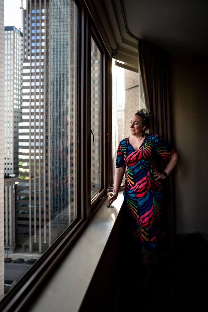 A white woman in a rainbow zebra print dress looking out of a large hotel window.