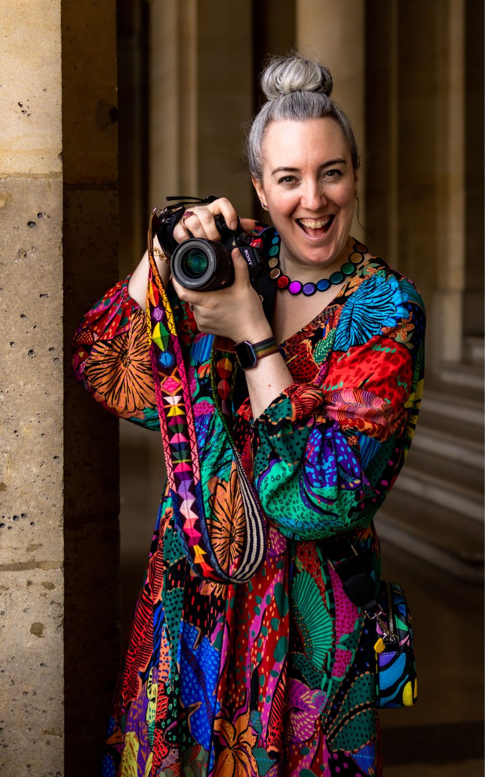 A happy, smiling white woman in a multi-coloured dress is holding her camera up to take photos in Paris