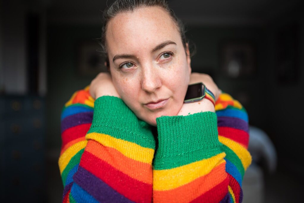 White woman wearing a rainbow striped jumper, looking pensive whilst hugging herself