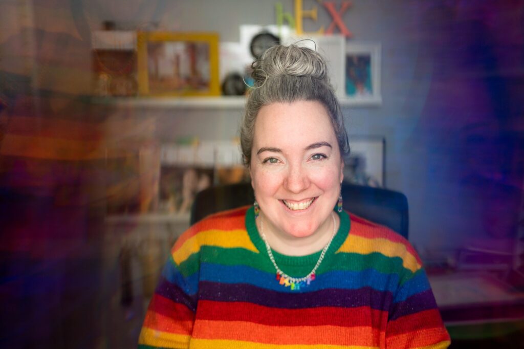 White woman wearing a rainbow striped jumper, looking happy in her office