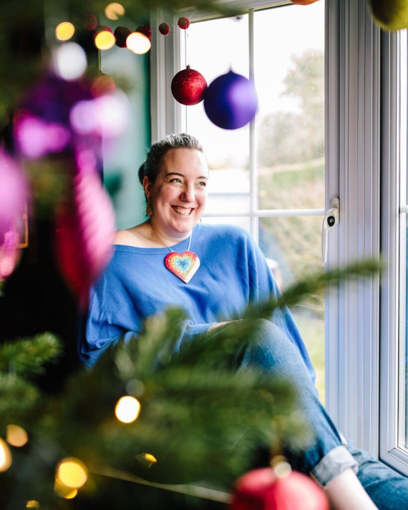White woman posing in a window at Christmas, surrounded by colourful baubles and lights, she's smiling and wearing a blue jumper and a large rainbow necklace.