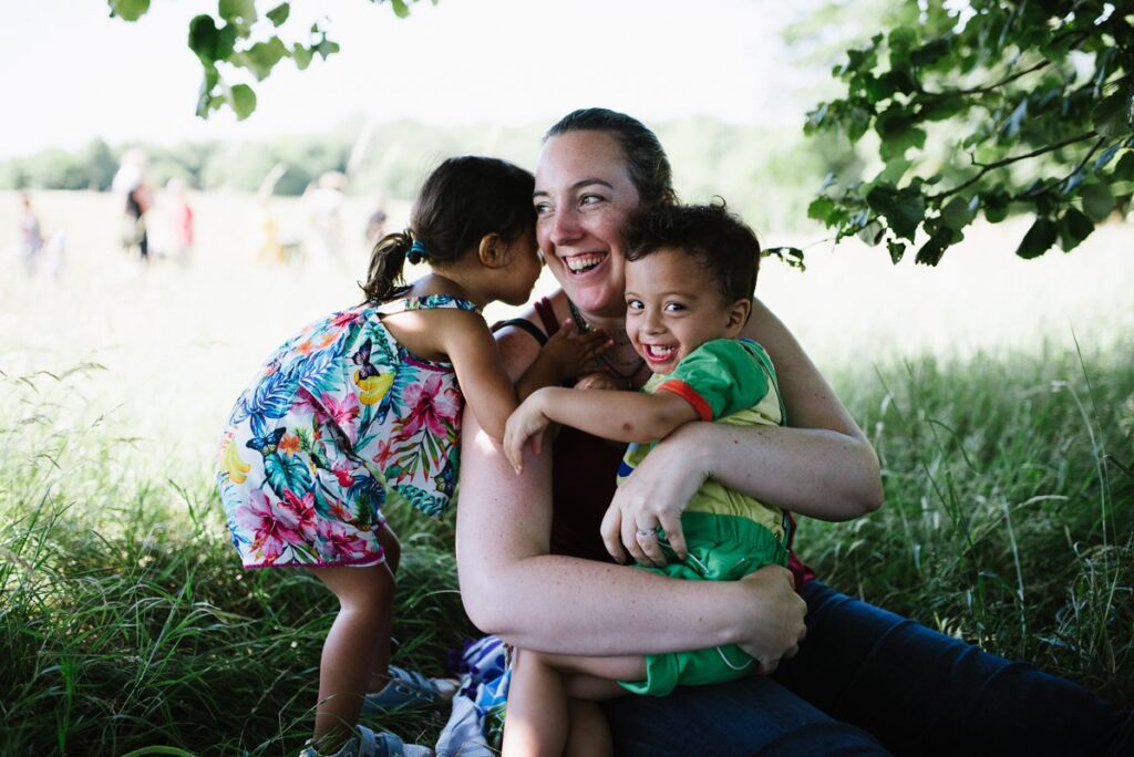 White mother cuddling her mixed-race twin children in a park, everyone looks happy.