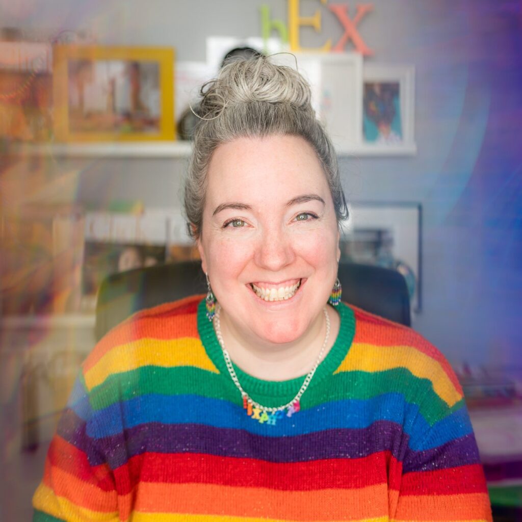 White woman wearing a rainbow striped jumper, looking happy in her office