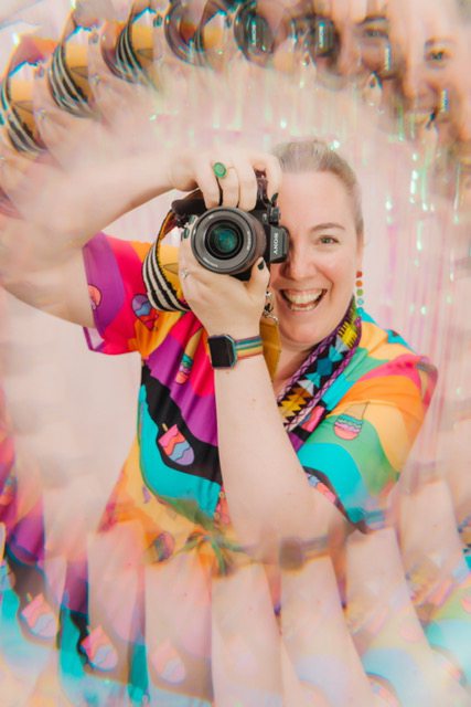 A white female photographer holding her camera up to her face with a big smile. She is wearing a rainbow coloured dress and is surrounded by a prism effect showing multiple reflections of herself.