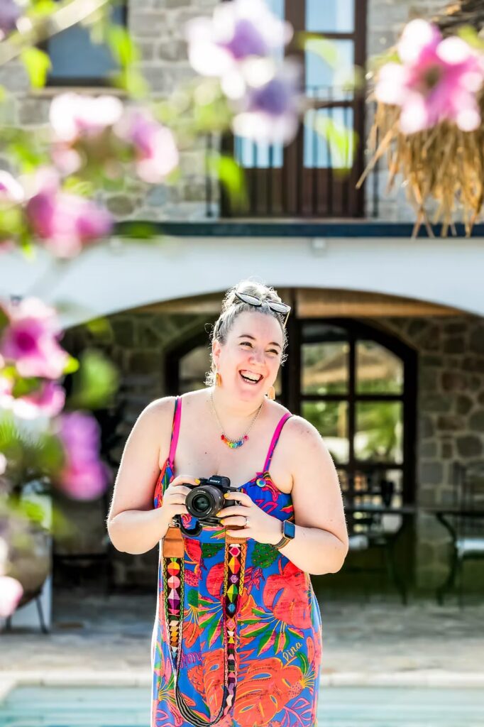 A white woman with grey hair laughing in the sunshine in Spain. She is holding a camera and wearing a blue and red jumpsuit.
