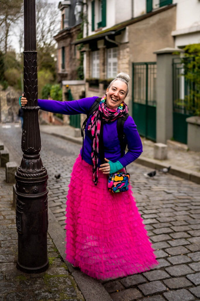 A happy white woman with silver hair is swinging joyfully from a lamppost in Paris. She is wearing a bright pink frilly skirt, purple jumper, pink patterned scarf and turquoise gloves and a rainbow pattern bag.