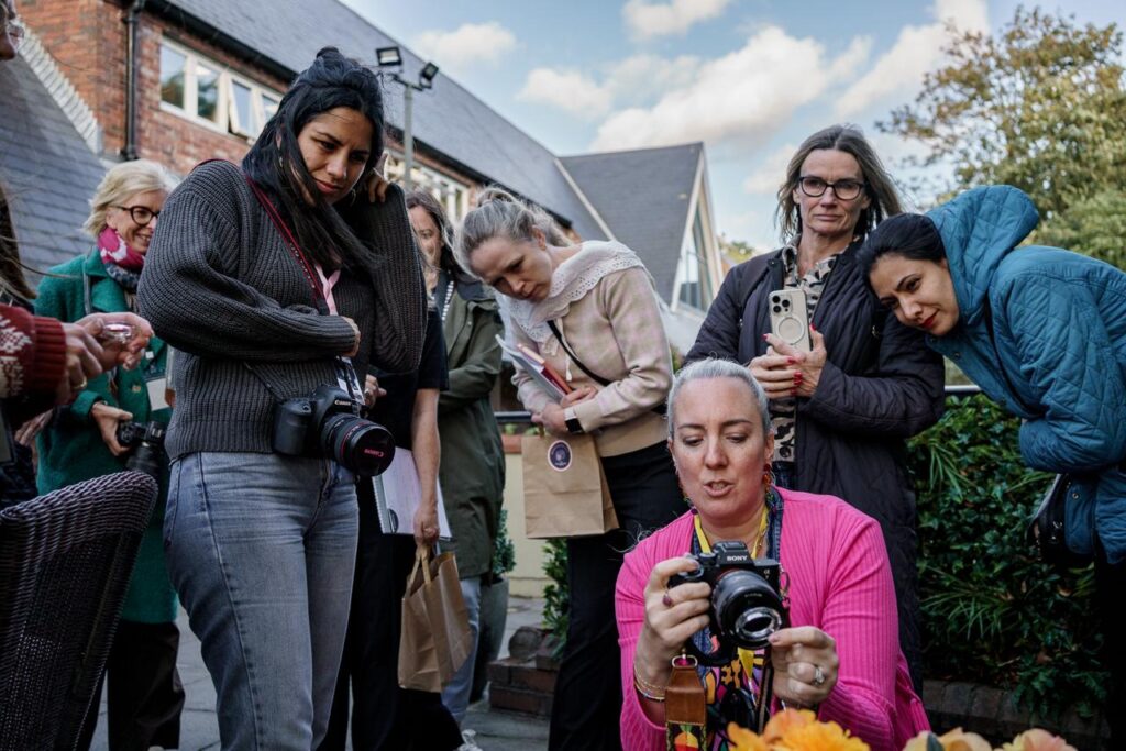 A white female photographer wearing a bright pink cardigan demonstrating a technique to her female students who surround her.