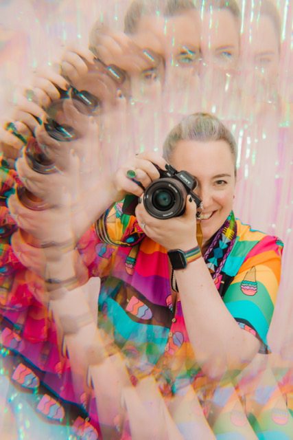 A white female photographer is holding her camera up to her eye and smiling from behind the lens, she is wearing a bright rainbow striped dress and has reflections of her circling around her through a ring prism.