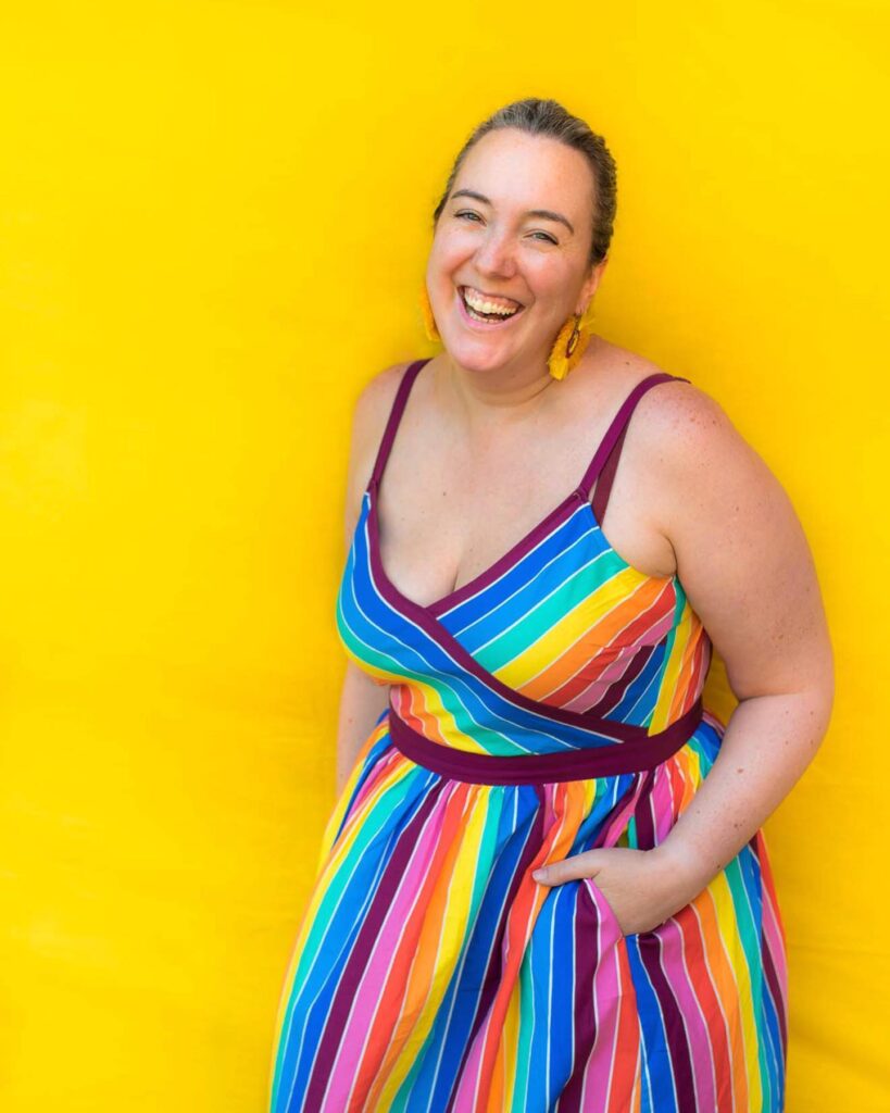 A happy white woman with brown hair is posing in front of a yellow backdrop and smiling, She is wearing a rainbow striped strappy dress and yellow earrings and has her hands in her pockets.