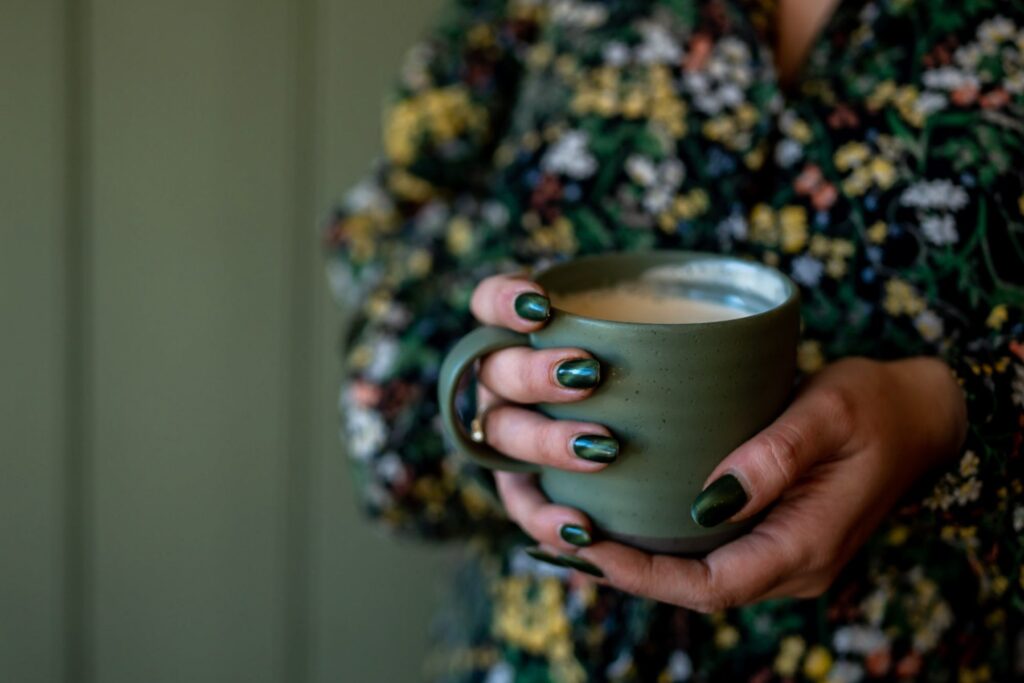 Close-up of a woman's hands holding a green mug of coffee with green painted fingernails, wearing a floral green dress and a green wall behind