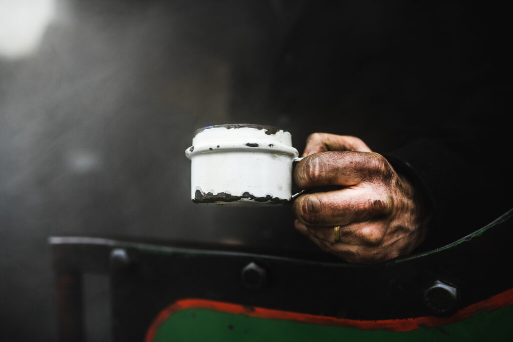A blackened dirty hand of a steam train worker holding a battered enamel mug of tea.