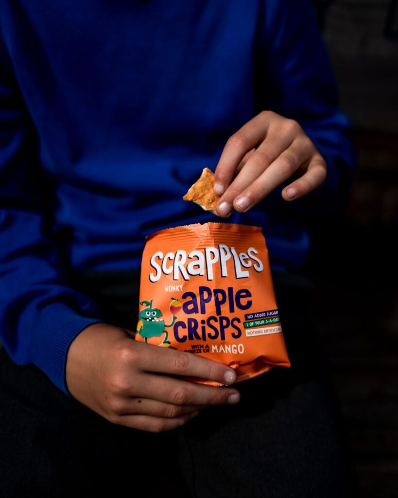 Close-up product photograph of a bag of Scrapples apple and mango crisps in the hands of a child who has taken on crisp out, with his blue school sweater behind.