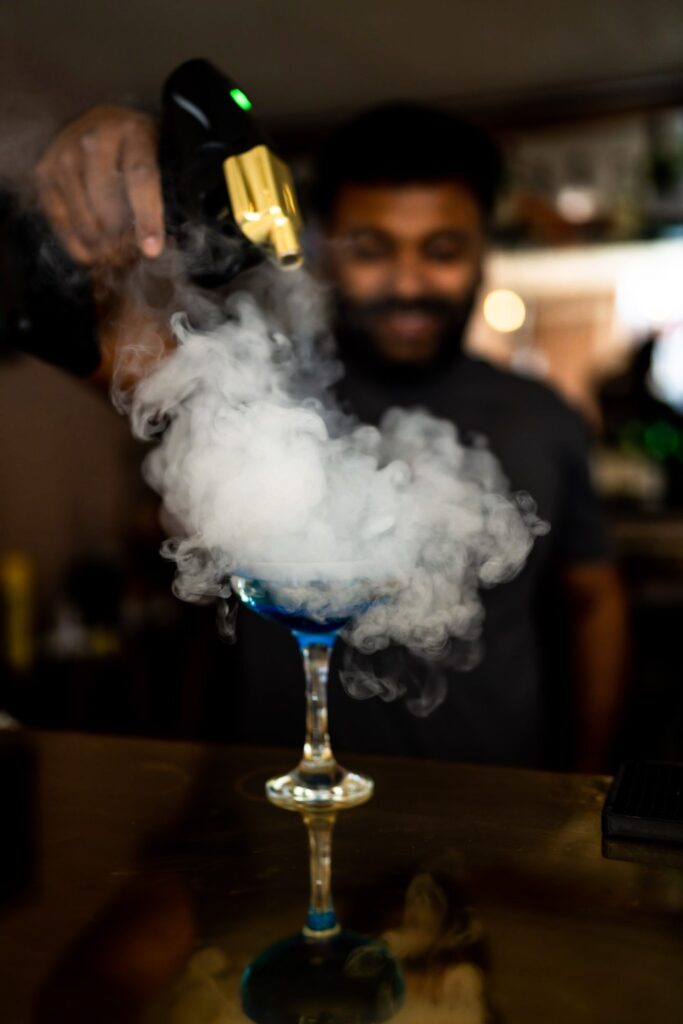 A smoky cloud enveloping a blue cocktail in a martini glass with the barman smiling in the background.