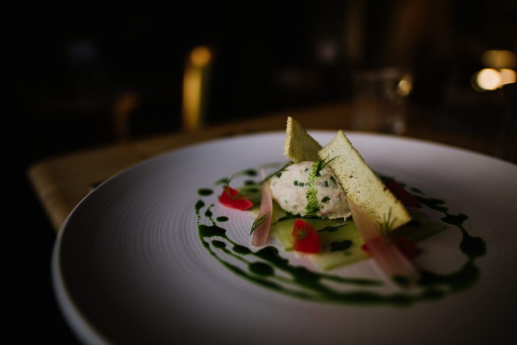 Beautifully decorated food on a white plate with greens and pinks dotted around and a dark moody background which gives a cosy vibe