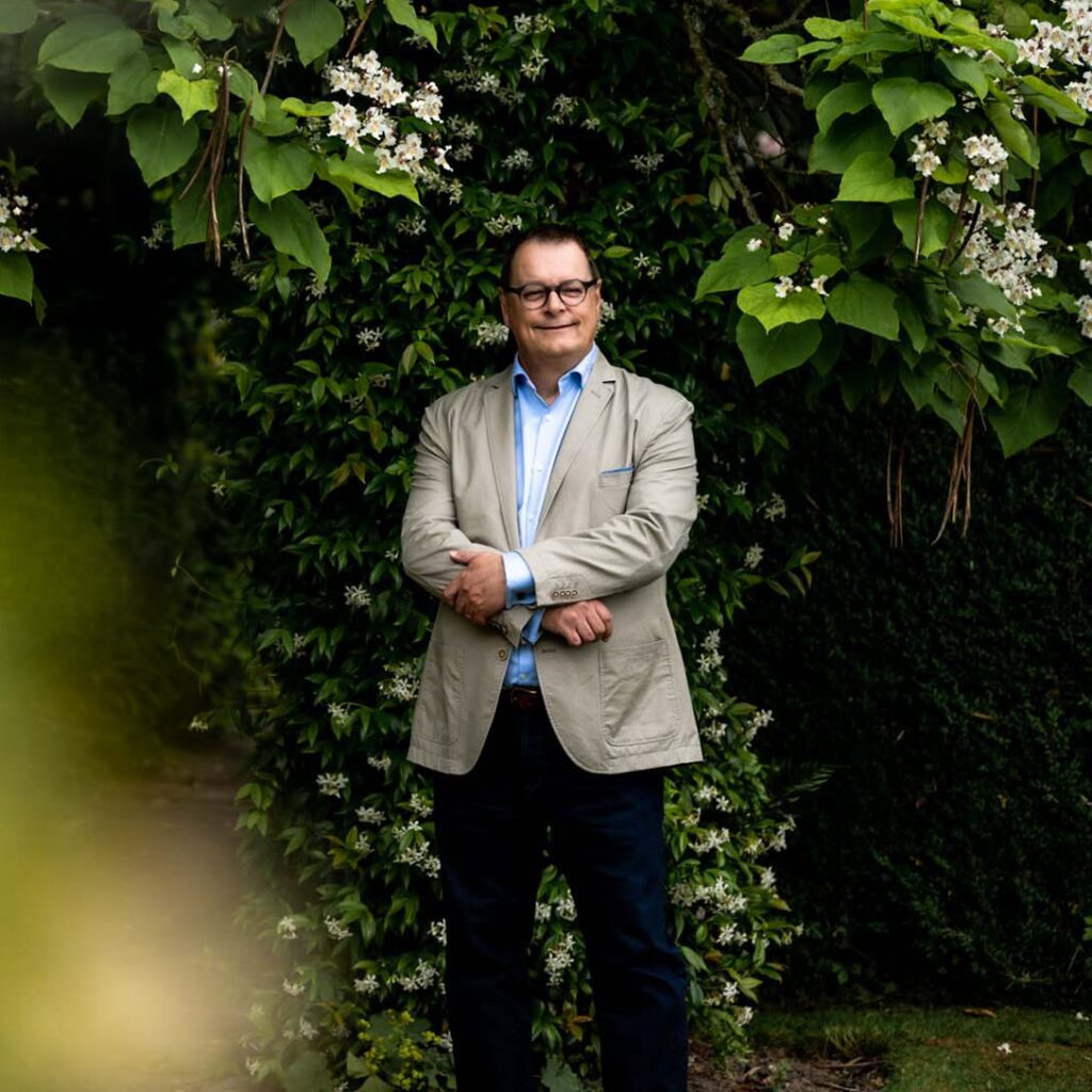 White man with a beige blazer and blue shirt standing surrounded by greenery in an outdoor location