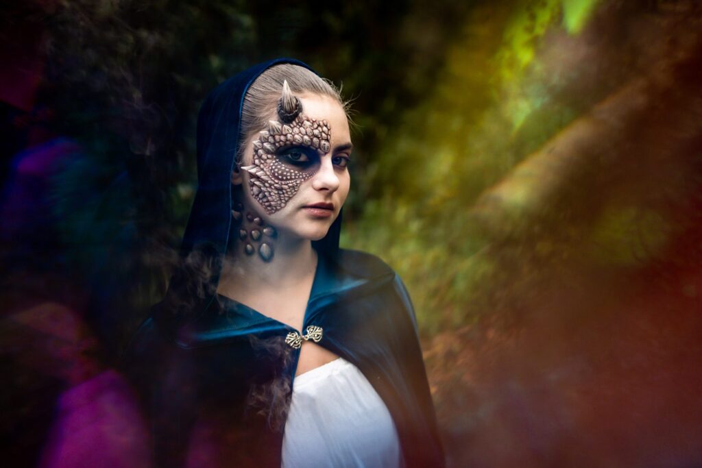 A young woman in a dark hooded cloak with intricate textured face paint across half her face by make-up artist Emese Csoma, lit by green and purple prism light in Buxted Park woods.