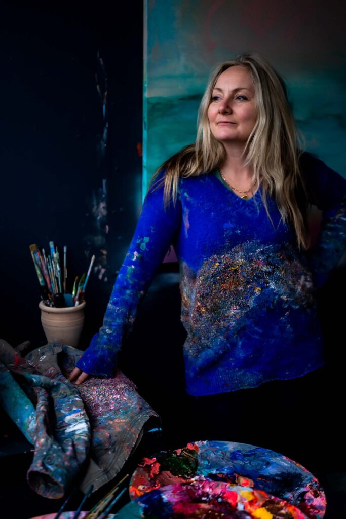 A white female artist with blonde hair in her colourful art studio. She is wearing a blue jumper covered in paint and posing by her paintings, brushes and palettes.