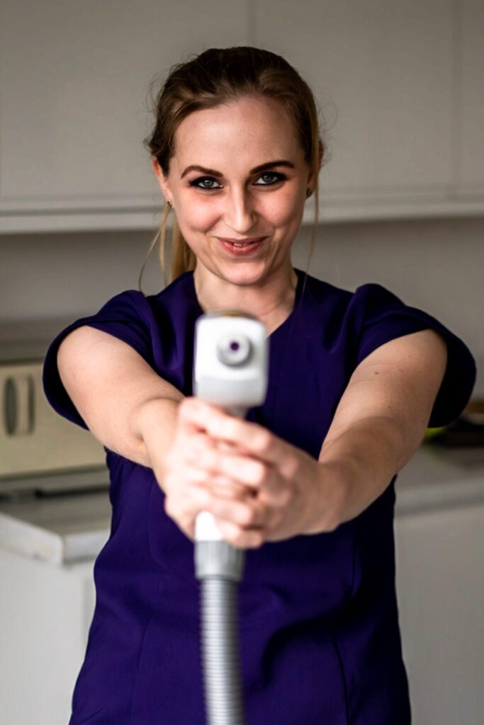 A white women with blonde hair tied up is holding a laser gun pointed at the camera like a playful Lara Croft with a half smile on her face, trying not to laugh.