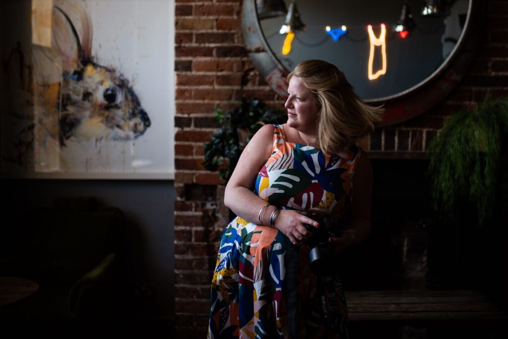 A blonde white woman swishes her hair in a quirky hotel. She is wearing a tropical print Boden dress.