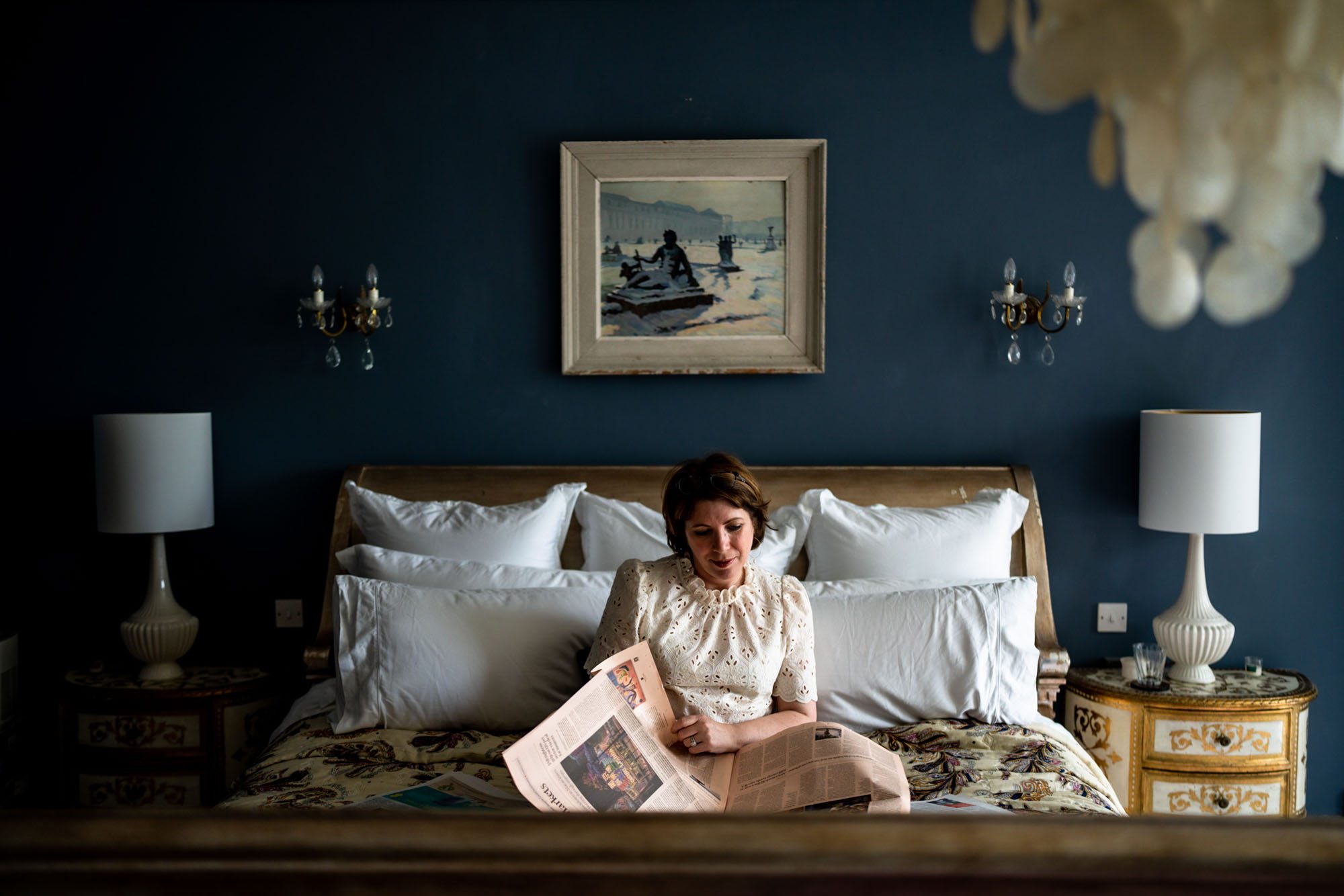 White woman with brown hair reading newspapers on a king-size bed against a navy blue wall with antique décor.