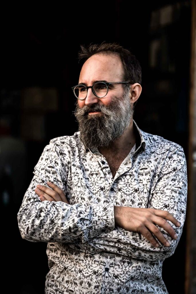 White man with brown hair and a greying beard, wearing glasses and a patterned white shirt with his arms folded, looking content off to the side with a dark background.