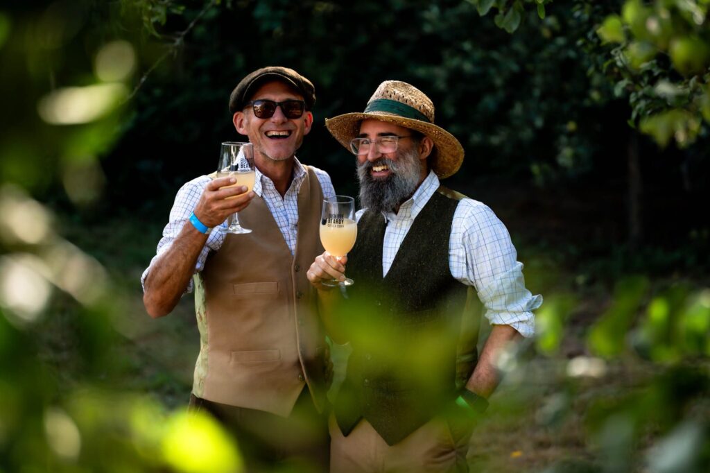 Two white men wearing jaunty English hats and waistcoats holding up glasses of cider in the sunshine, surrounded by the foliage of the orchard where it was made. They are happy, laughing and one of them is winking.