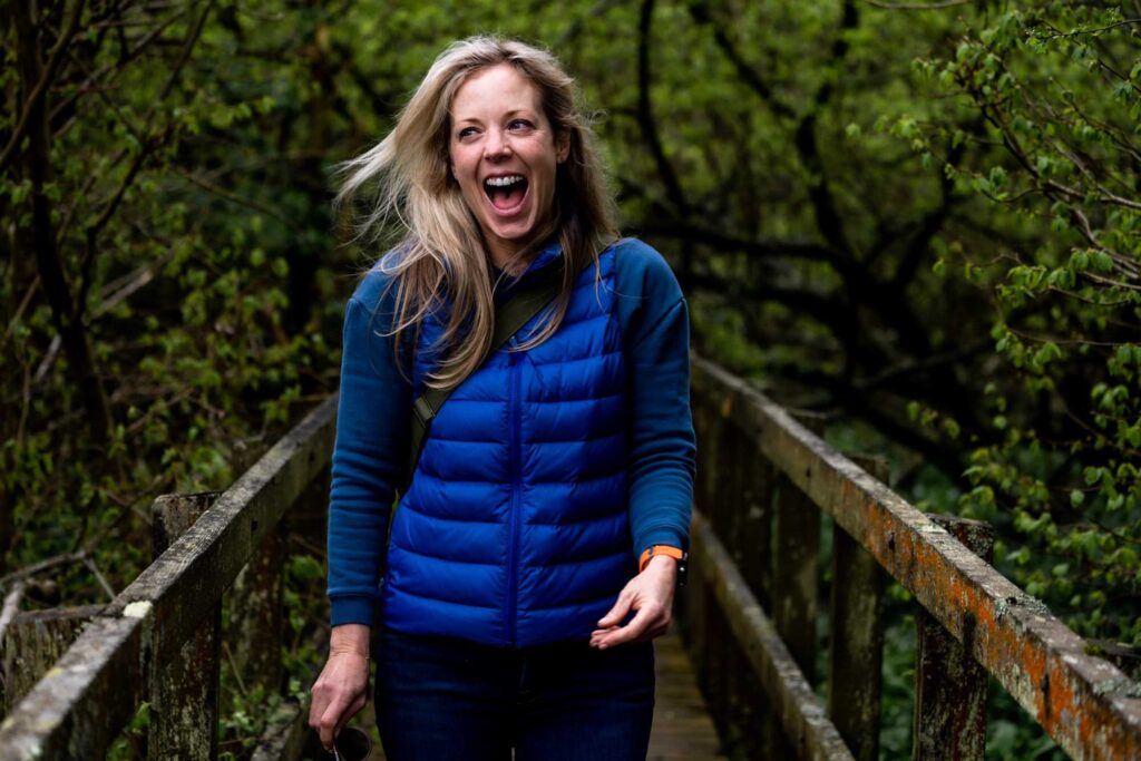 White woman with blond hair wearing a blue gilet on a wooden bridge in the woods, she's laughing and looks very happy