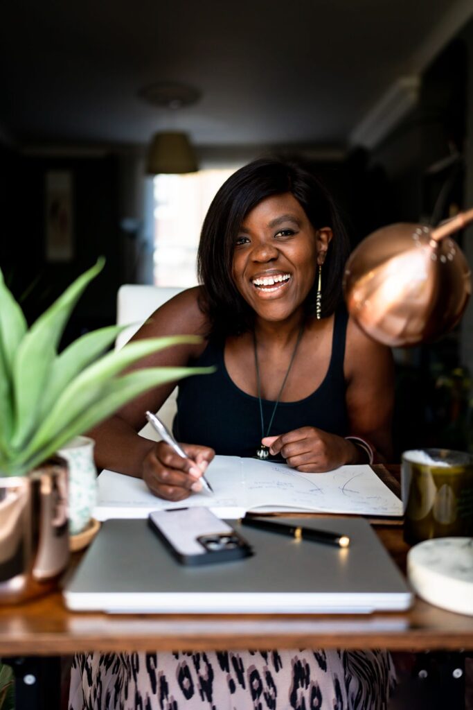 A black woman in a black wig sitting writing notes at her desk, smiling happily at the camera.