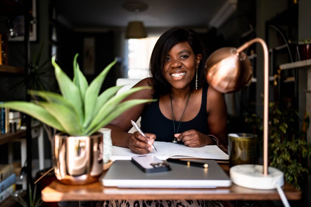 A black woman in a black wig sitting writing notes at her desk, smiling happily at the camera.