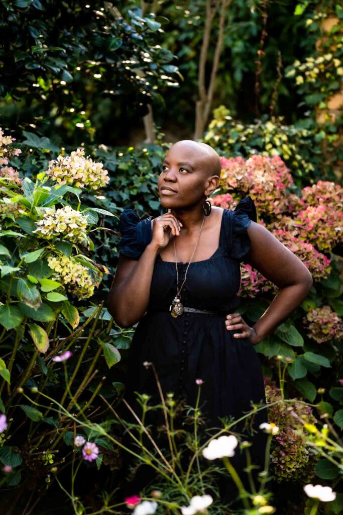 A bald black woman posing with her hand under her chin, looking pensive, among her hydrangeas in her garden