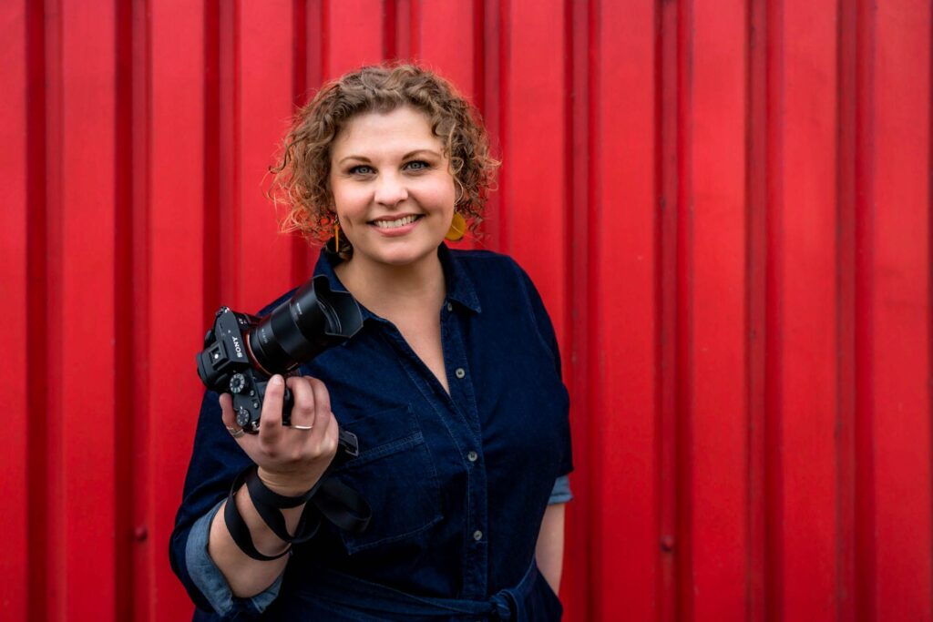 A white female photographer with light brown curly hair poses in a denim dress for a headshot with a red backdrop