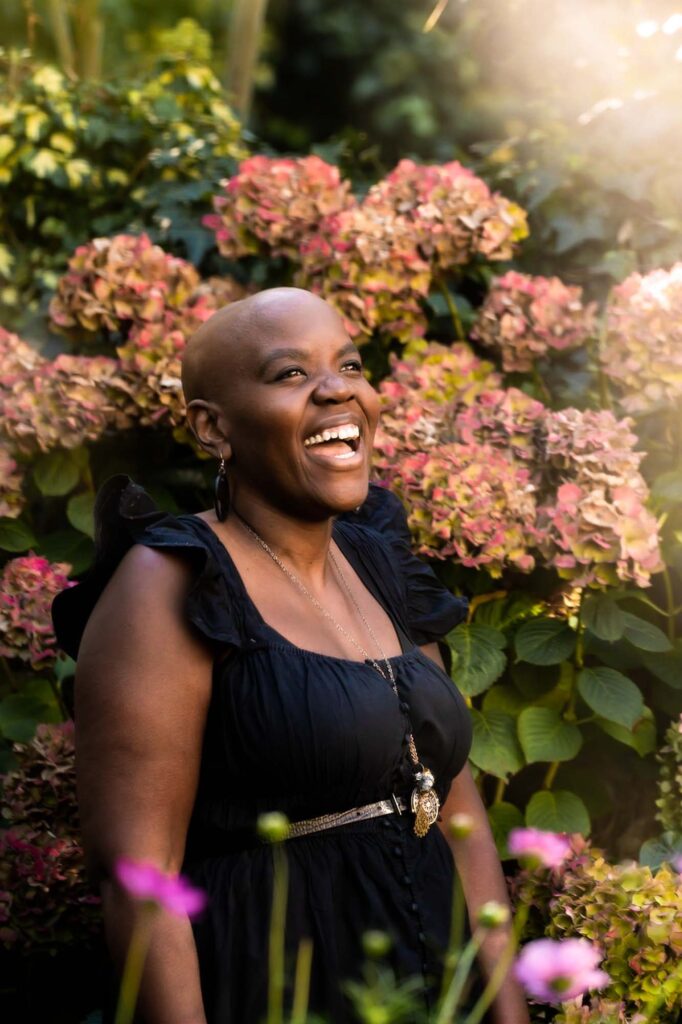 A bald black woman laughing joyfully in her garden with the sunshine bursting from the corner and big hydrangeas behind her.