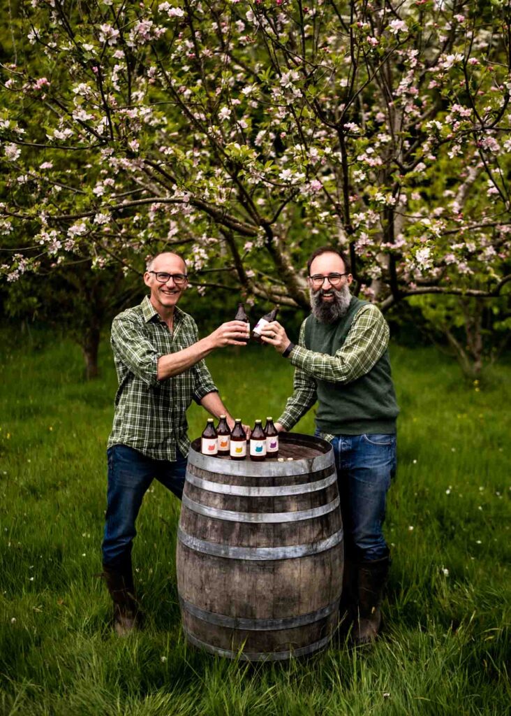 Steve and Phil AKA Bignose & Beardy, 2 white men are clinking glasses of cider, surrounded by blossoming apple trees in their orchard in East Sussex.