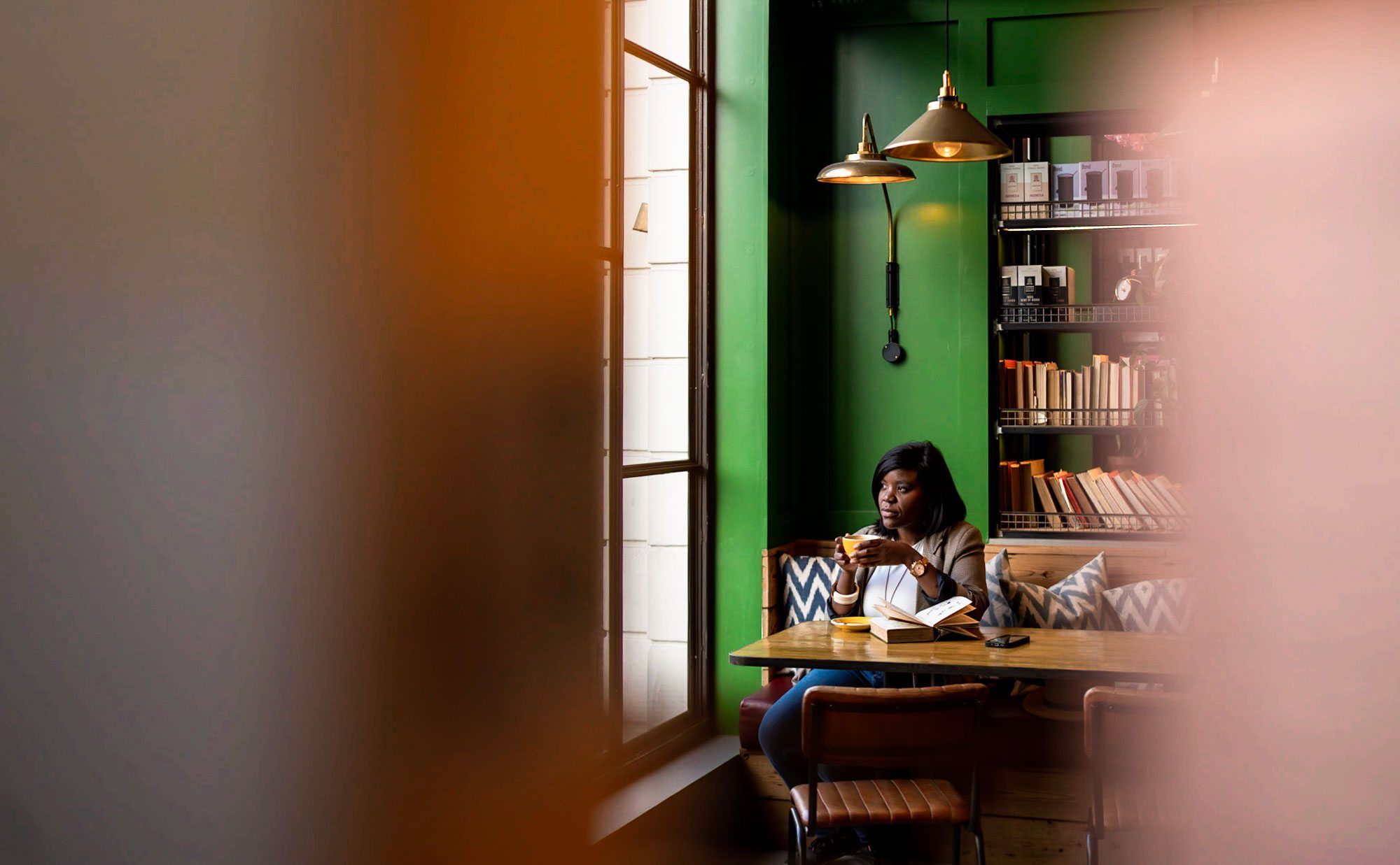 A Black woman in a taupe blazer and blue jeans, sitting with a coffee in a London café with a green wall, framed between books on a shelf with another full bookshelf behind her.