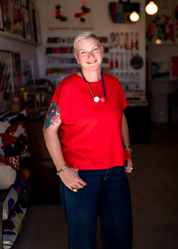 A white woman with cropped blonde hair and tattoos wearing a red t-shirt.
