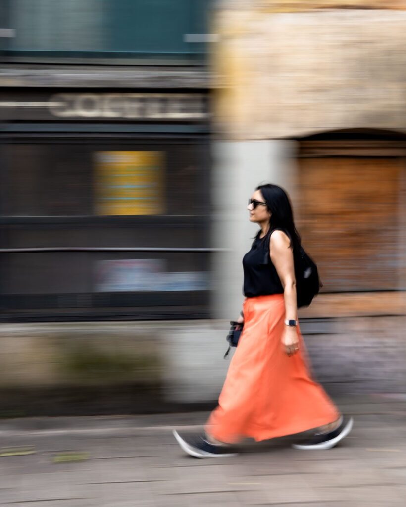 An Asian woman in an orange maxi skirt and black vest top, walking fast in a blur through London