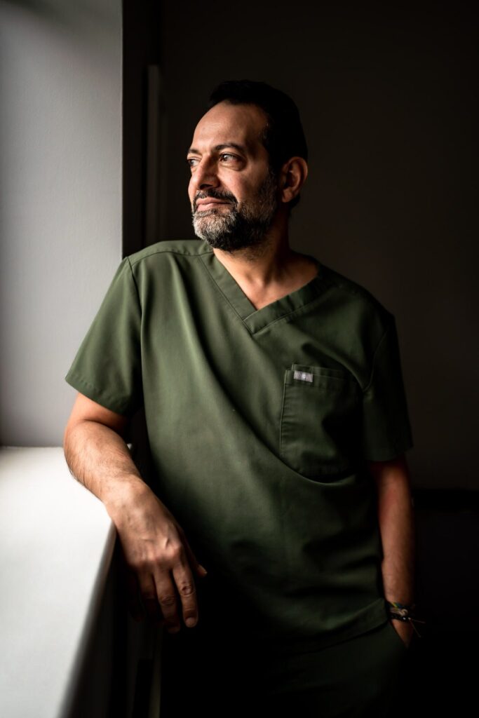 Turkish male dentist looking out of the window in his surgery, wearing dark green scrubs and looking relaxed.
