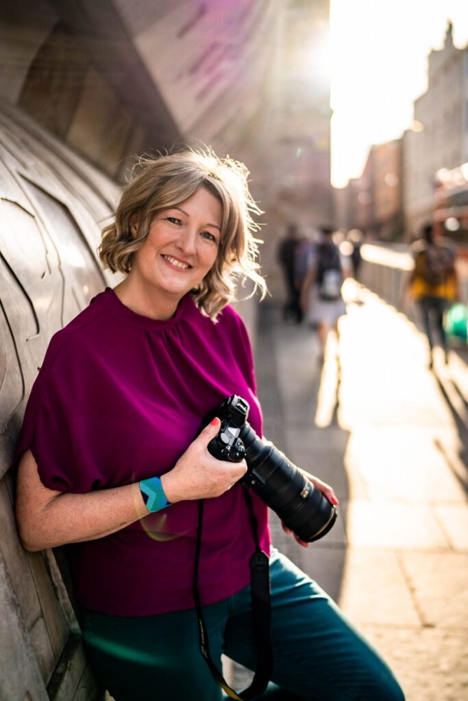 White woman with curly blonde hair holding a camera on The Royal Mile in Edinburgh with the sun shining behind her. She is smiling and wearing a magenta pink top.